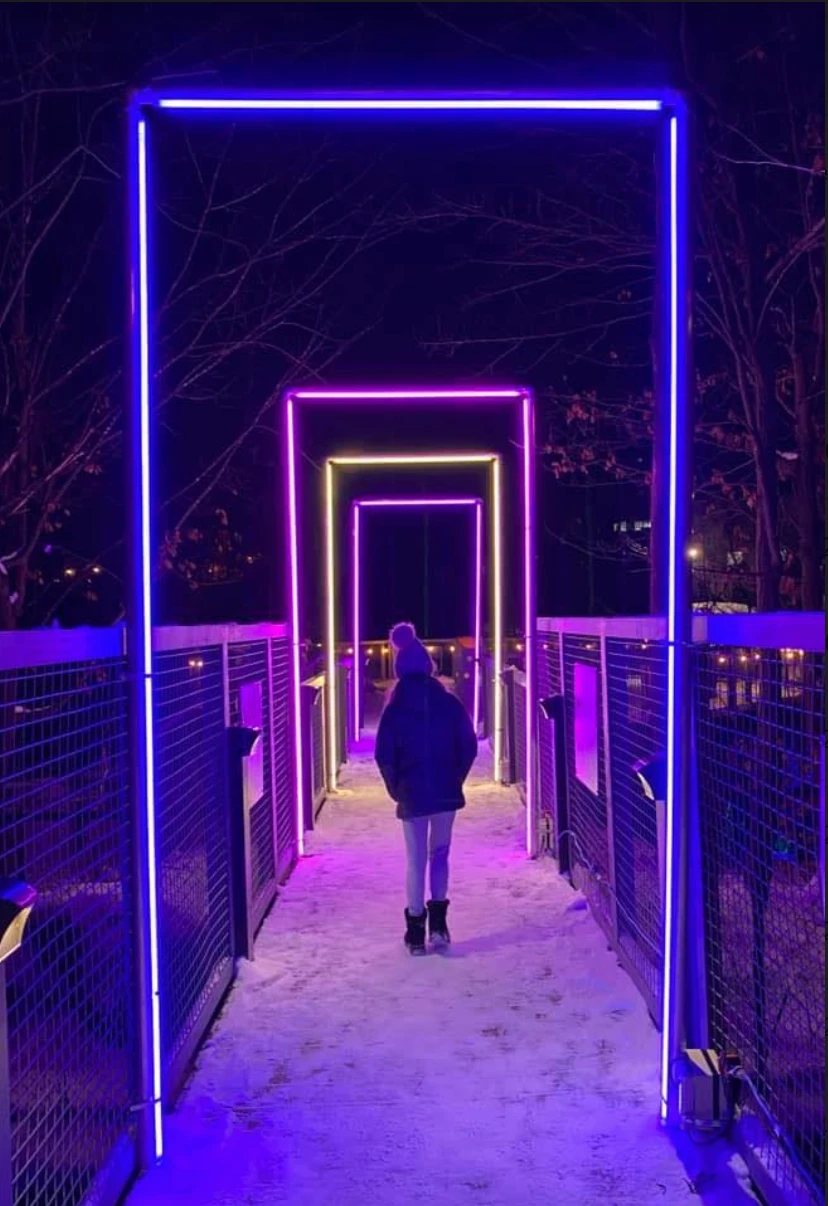 Capture-decran-le-2023-02-01-a-08.33.57 Un chemin de nuit orné d'une série de frames lumineuses bleues, violettes et jaunes, bordé par une barrière métallique et recouvert de neige. Un individu marche dans ce décor coloré.