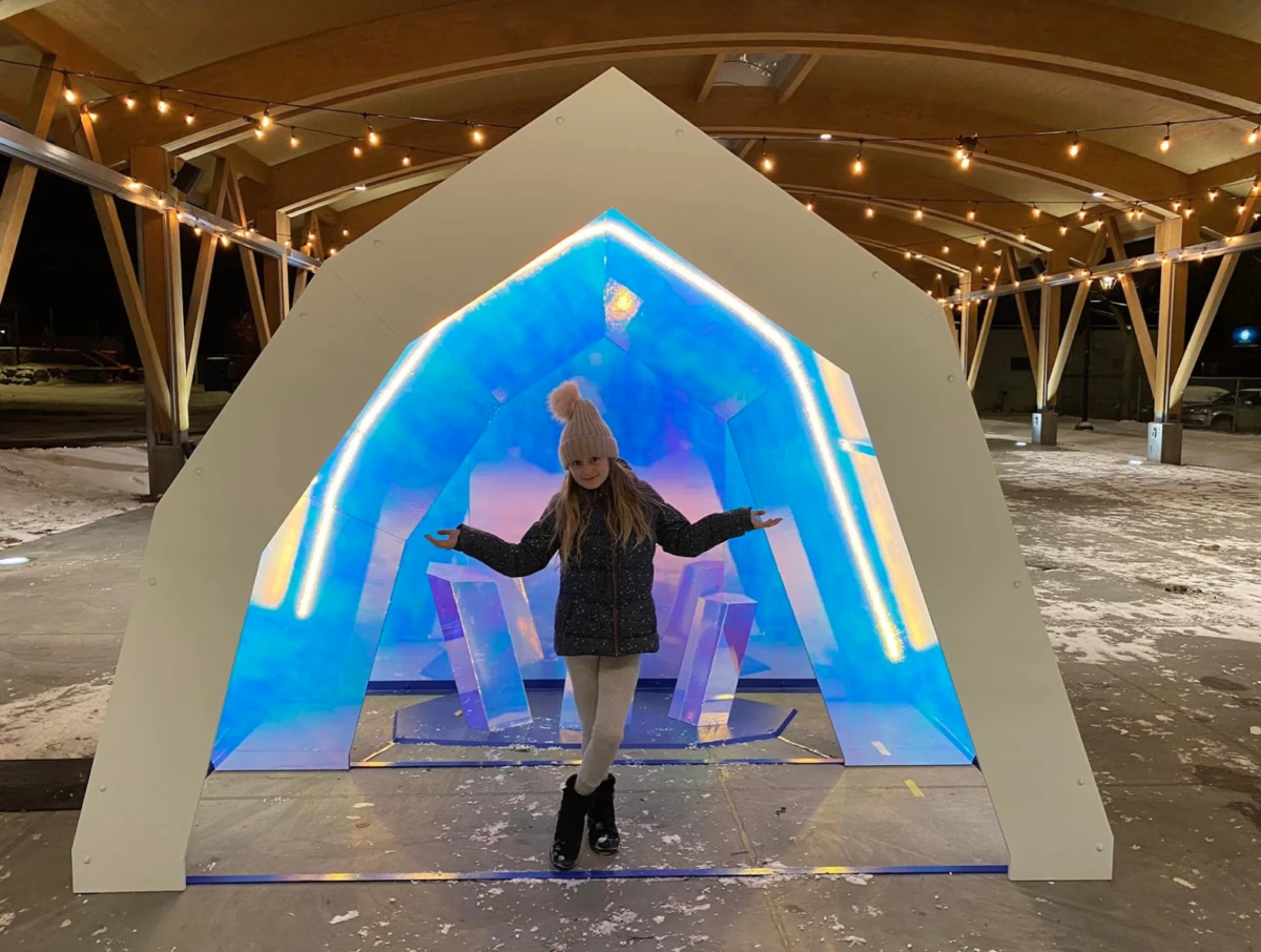Capture-decran-le-2023-02-01-a-08.34.05-e1744219723166 Une jeune femme pose devant une structure lumineuse bleue, avec des lumières décoratives dans le ciel nocturne, symbolisant la magie de Noël.